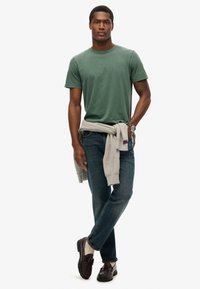 T-shirt en coton vert, sweat-shirt gris clair noué autour de la taille, jeans en denim bleu, mocassins marron avec semelles noires, cheveux mi-longs.