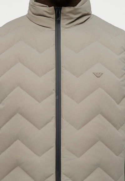 Μπεζ μπουφάν με επένδυση σε σχήμα βέλους, μαύρο κεντρικό φερμουάρ και μικρό λογότυπο αετού στο αριστερό στήθος.