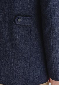 Morska niebieska wełniana kurtka, rękaw ukazujący teksturowany materiał, mała kieszonka z czarnym guzikiem i staranne przeszycie wzdłuż szwu.