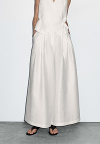 Vit linneärmlös topp med en omlottdesign och sidoband, tillsammans med vida vita linnebyxor. Svarta platta sandaler fulländar looken.