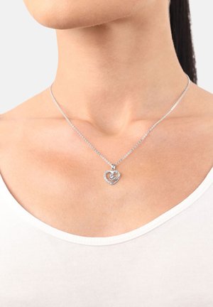 Collana con pendente a forma di cuore in argento con un piccolo gemma al centro, indossata intorno al collo di una donna sopra una maglietta bianca.