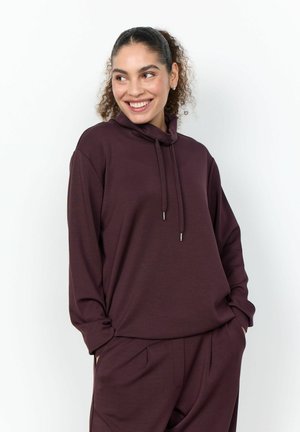 Giovane donna con capelli ricci raccolti in una coda di cavallo che indossa una felpa con cappuccio bordeaux scuro e pantaloni abbinati, mani nelle tasche, sorridente e guarda di lato.