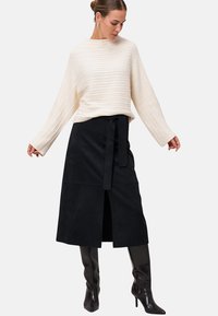 Femme portant un pull crème en maille torsadée, une jupe midi noire ceinturée avec fente devant, et des bottes noires à talons montant jusqu'au genou, posant les bras légèrement écartés.