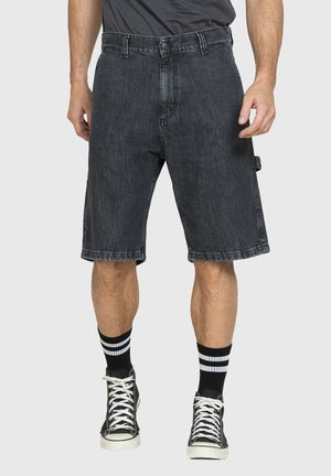 Uomo che indossa pantaloncini di jeans neri, calze a righe bianche e nere e scarpe da ginnastica nere alte, in piedi contro uno sfondo semplice.