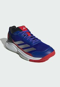 Chaussure de sport bleue avec accents rouges et gris. Fabriquée en mesh et matériaux synthétiques, avec une semelle texturée et trois bandes horizontales.