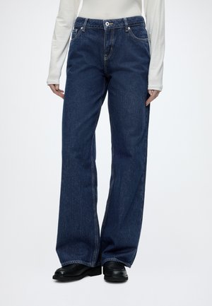 Personne portant un jean bleu foncé à jambes larges, des mocassins noirs et un haut blanc à manches longues, debout devant un fond uni.