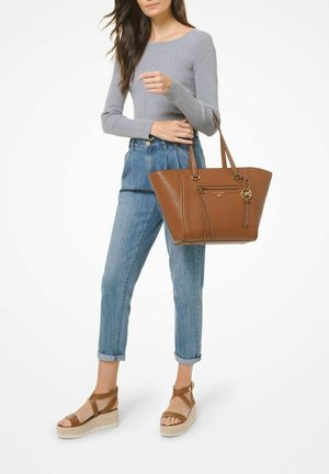 Vrouw draagt een grijs shirt met lange mouwen, opgerolde blauwe spijkerbroek en bruine sleehakken, en houdt een grote bruine leren shopper met gouden accenten vast.