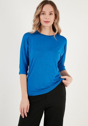 Blue, lightweight, long-sleeve top with a round neckline and 3/4 sleeves, featuring a smooth texture and relaxed fit. Paired with black trousers.