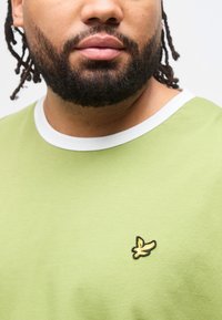 Άνδρας με γενειάδα που φοράει ανοιχτοπράσινο π ουκάμισο με άσπρο γιακά και ένα μικρό κίτρινο πουλί στο στήθος, σε λευκό φόντο.