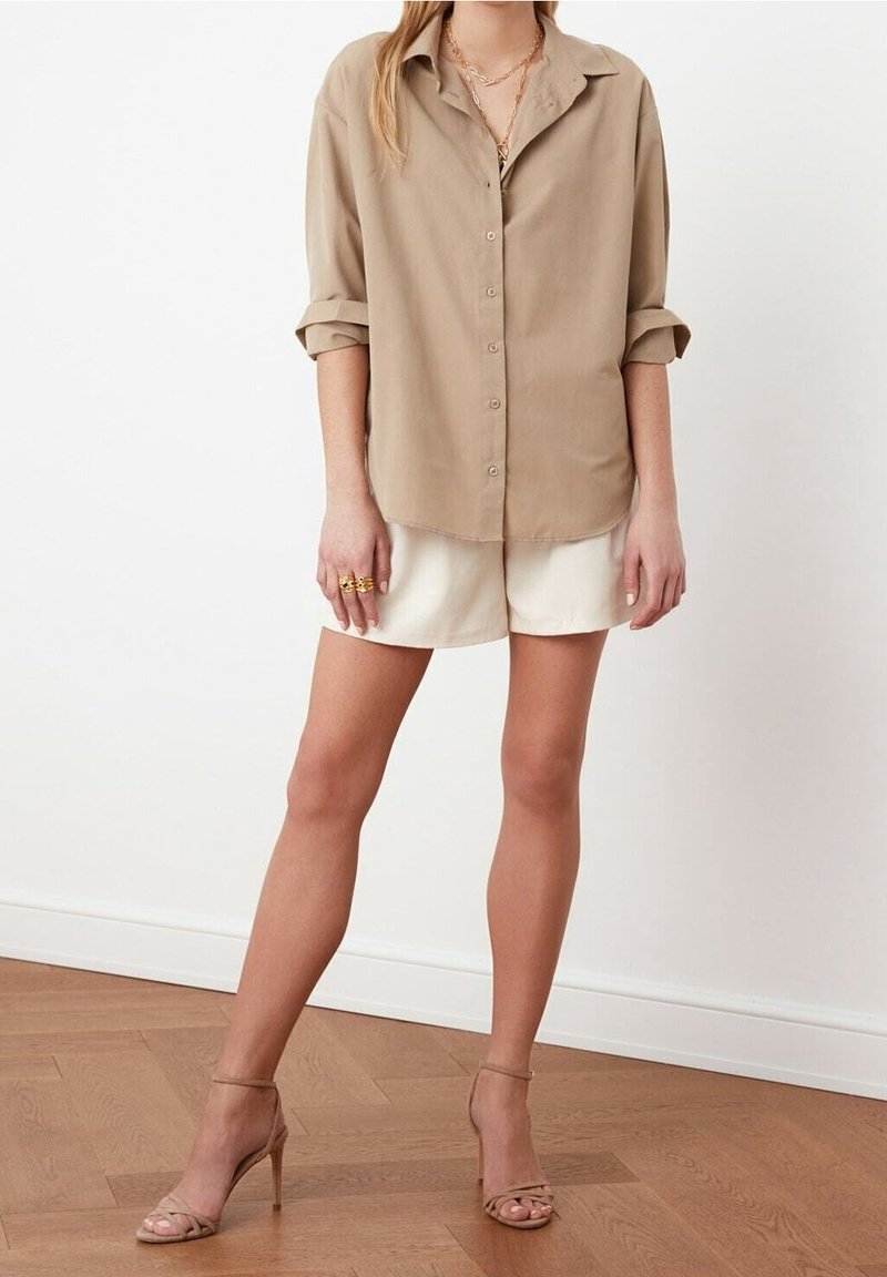 Personne portant une chemise beige boutonnée avec manches retroussées, un short blanc, des bijoux en or et des sandales à talons nus, debout sur un sol en bois.