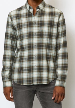 Man draagt een overhemd met lange mouwen in olijfgroen, wit en lichtblauw ruitpatroon, gecombineerd met donkergrijze spijkerbroek, voor een effen achtergrond.
