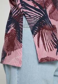 Różowa bluzka z krótkim rękawem z głębokimi granatowymi i burgundowymi wzorami liści, z bocznymi rozcięciami i luźnym krojem, zestawiona z jasnoniebieskimi dżinsami.