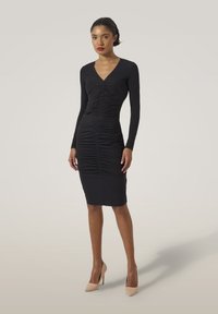 Svart, figurnära klänning med långa ärmar, v-ringning och rynkdetaljer. Har en knälång fåll och en mjuk, elastisk tygkvalitet.