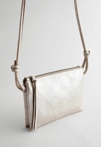Metalliczna srebrna torba crossbody wykonana ze skóry. Posiada zamykane wieko, płaski kształt i szczegół w postaci węzła na pasku, co umożliwia regulację długości.