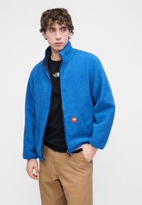 Μπλε φλις μπουφάν με φερμουάρ μπροστά, ψηλό γιακά και πλαϊνές τσέπες. Διαθέτει υφή υφάσματος και μικρό λογότυπο στη θήκη του στήθους.