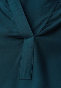 Blusa teal in tessuto dalla texture liscia; presenta un design a V e un dettaglio centrale della cucitura. Linee pulite e stile minimalista.