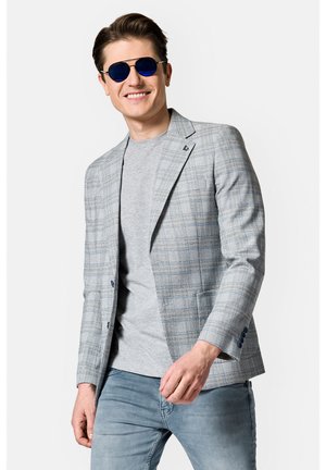 Jasnoszary blazer w kratę z niebieskimi akcentami, noszony na szarym t-shircie, w połączeniu z jasnymi dżinsami. Model nosi niebieskie okulary przeciwsłoneczne.