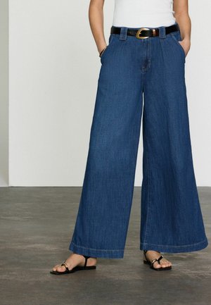 Tumedast sinisest denimpüksid, laiad, kõrge vöökoha ja esipocketitega, must vöö kuldse pandlaga. Mustad lamedad sandaalid.