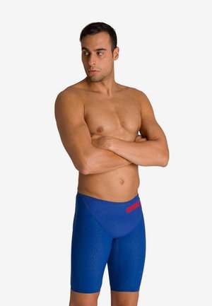 Man met gekruiste armen, gekleed in blauwe wedstrijdbadkleding met een rood merklogo, op een witte achtergrond.