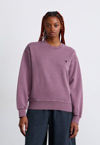 Young woman with long red braids wearing a loose, faded purple sweatshirt and dark pants, standing against a plain white background.