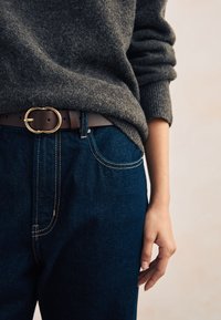 Pull en tricot gris, jean denim bleu foncé avec des coutures jaunes contrastantes, et une ceinture marron avec une boucle circulaire dorée.