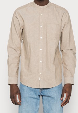 Homme portant une chemise beige à manches longues avec col mao et poche poitrine, associée à un jean bleu clair.