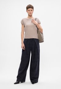 Beżowa bluzka z krótkim rękawem, szerokie spodnie w ciemną kratę i czarne buty. Niesie szaro-skalną torbę. Prosty, czysty design.