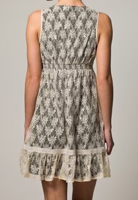 Pitkä pitsimekko, jossa on rypytetty vyötärö, beige ja kerma väreissä kukalliset kuviot sekä röyhelöhelma. Takakatsaus näyttää ilman hihoja -muotoilun.