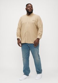 Beige színű, hosszú ujjú, slim fit gombos ing, világoskék farmernadrággal és fehér sportcipővel párosítva. Az ingen kis logó található.