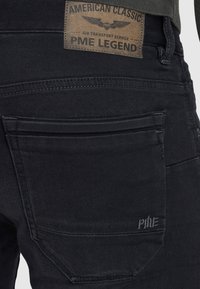 Gros plan de la poche arrière d'un jean en denim noir avec une étiquette en cuir marron indiquant "American Classic PME Legend" et un petit logo PME brodé.