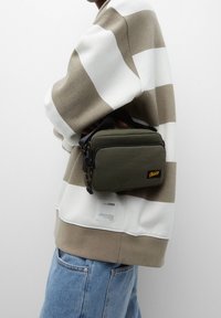 Sac rectangulaire vert avec une fermeture éclair noire, surface texturée, logo jaune et accents noirs, tenu sous un pull rayé marron et blanc.