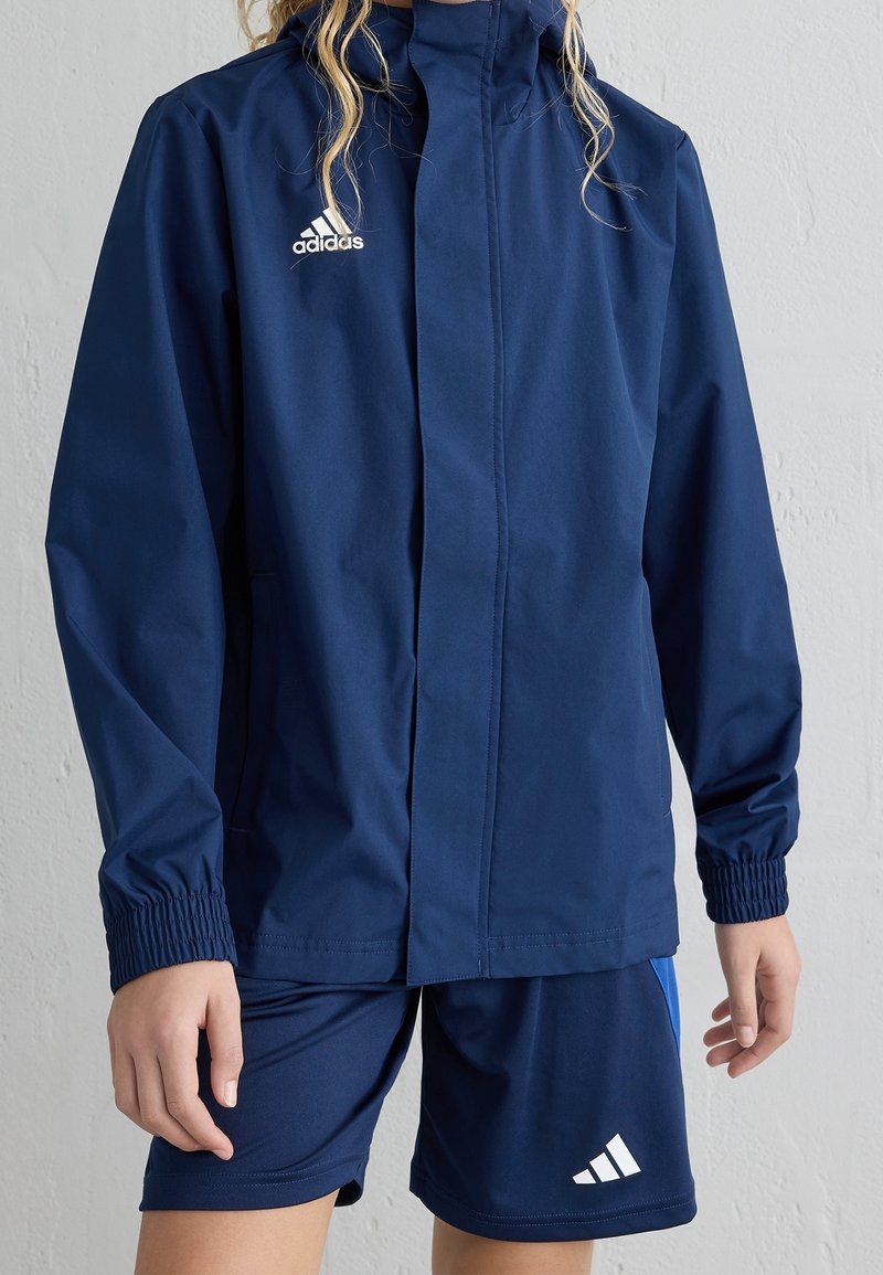 Marinblå sportjacka med hög krage och elastiska ärmslut. Har dragkedja fram, lättviktsmaterial och vit Adidas-logga.