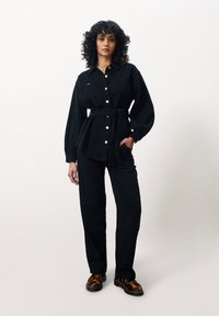 Chemise en jean noir à manches longues, avec des boutons à l'avant et une ceinture à nouer, assortie à un pantalon en jean noir et des sabots à motifs.