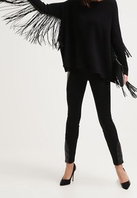 Kvinna i svart fransad tröja, svarta tajta byxor med läderlappar och svarta högklackade skor, som håller en fransad kuvertväska.