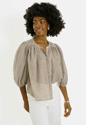 Femme avec coiffure afro portant des boucles d'oreilles argentées, un chemisier beige et blanc rayé avec des manches bouffantes et un pantalon blanc, souriante et regardant sur le côté.