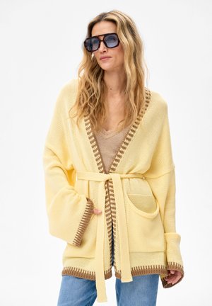 Kobieta o długich, falowanych blond włosach, nosząca duże okulary przeciwsłoneczne, żółty kardigan z paskiem i brązowymi wykończeniami, beżową bluzkę oraz niebieskie dżinsy.