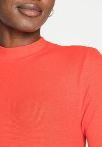 Röd ribbad topp med hög hals och en slät textur, med en figurnära design som framhäver halslinjen och axelområdena.