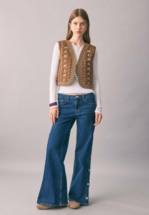 Rjav kvačkast telovnik z izvezenimi motivi, nošen čez belo srajco z dolgimi rokavi, v kombinaciji z moderimi širokimi jeans hlačami, ki imajo gumbne detajle.