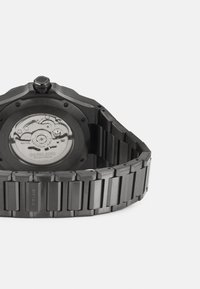 Montre en acier inoxydable noir avec un boîtier octogonal, un bracelet texturé et un fond de boîtier transparent affichant le mouvement mécanique.