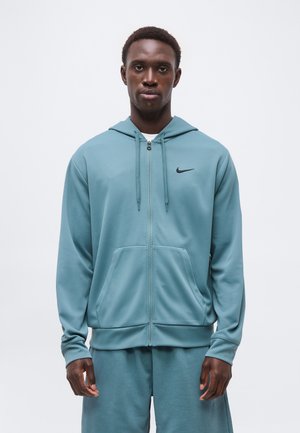 Jaunas vyras vilki šviesiai mėlyną Nike su užtrauktuku megztinį su gobtuvu ir tinkamas šortus, stovi prieš baltą vienodą foną.