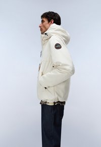 Mann in cremefarbener Winterjacke mit Kapuze und Aufnäher am Ärmel, dunklen Jeans und kariertem Hemd darunter, der im Profil vor hellem Hintergrund steht.