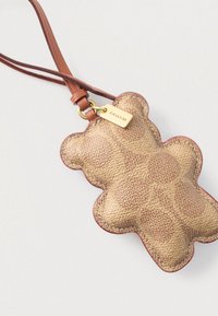 Barna bőr medve alakú kulcstartó dombornyomott logóval, texturált felülettel és kontrasztos piros szegéllyel. Levehető pánttal rendelkezik a sokoldalúság érdekében.