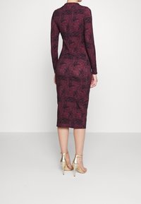 Vestido ajustado burdeos de mangas largas con un patrón floral, con un dobladillo a la altura de la rodilla y una textura suave. Se lleva con tacones de tiras doradas al tobillo.