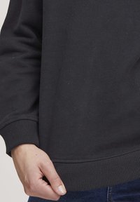 Felpa nera con polsini e orlo a costine, caratterizzata da una texture liscia. Una mano tiene il tessuto vicino al bordo inferiore, evidenziando la vestibilità.
