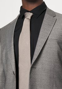 Šedý vlnený oblekový kabát s špičatým klopovým límcem, kombinovaný s béžovou kravatou s rybí kostrovým vzorem na černé košili. Viditelné detaily texturované tkaniny.