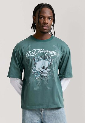 Joven con trenzas que lleva una camiseta verde de Ed Hardy con un gráfico de una calavera y rosas azules sobre una camisa blanca de manga larga.