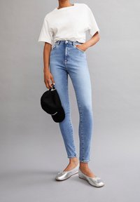 Höga ljusblå jeans, vit oversized t-shirt, silvriga ballerina skor och en svart keps som hålls i handen. Slätt tyg, figursydd design.