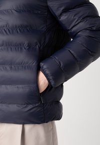 Giacca a vento blu navy con design a quadrati orizzontali, dotata di tasca con zip e polsini a coste. Tessuto liscio e leggero con una finitura lucida.