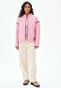 Žena s dlhými kučeravými vlasmi oblečená v ružovej prešívanej bunde, bielom topu, krémových širokých nohaviciach a béžových turistických topánkach stojí na bielom pozadí.