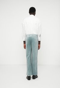 Calças em veludo cotelê azul-claro com pernas largas e uma textura suave, combinadas com uma camisa branca com riscas finas. A vista de trás mostra dois bolsos.